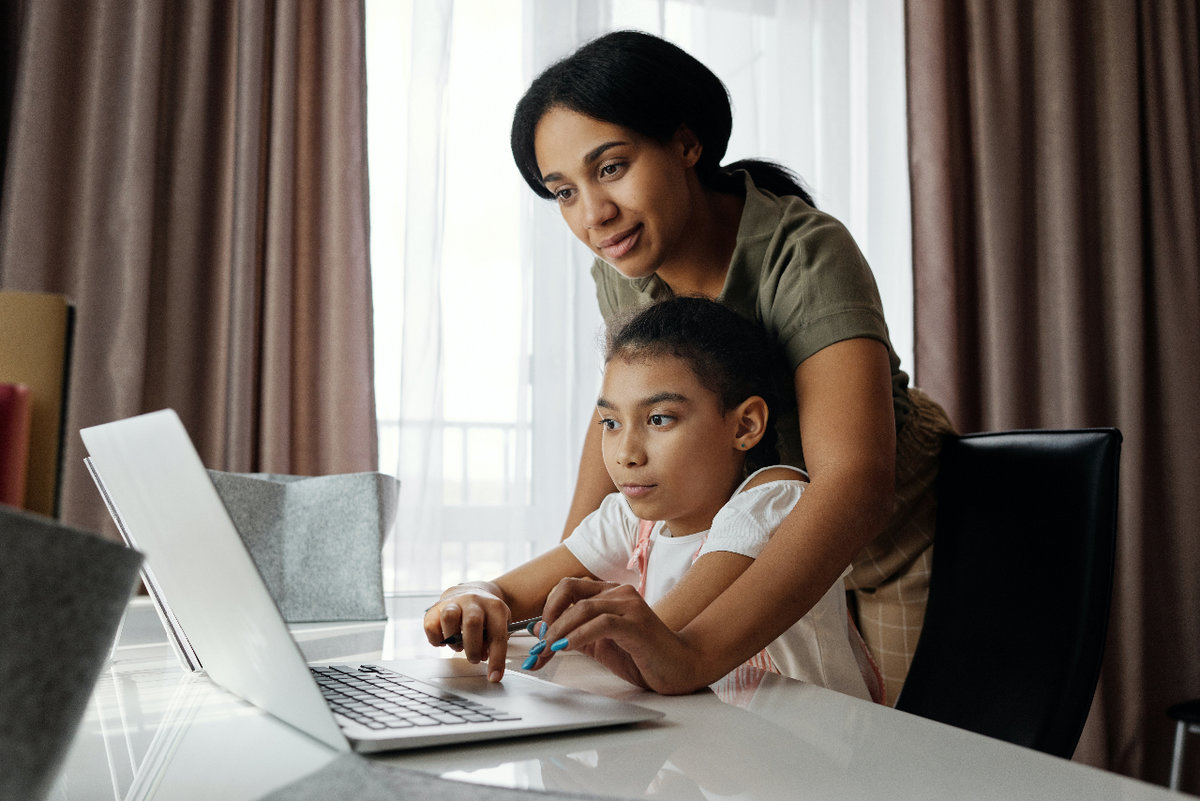 A mother stands over her child while she sits and works on a computer. The mother has her arms wrapped around her daughter, helping her with something on the laptop.